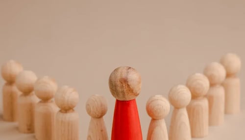 A single red figure stands prominently among a cluster of wooden figures, emphasizing its unique presence