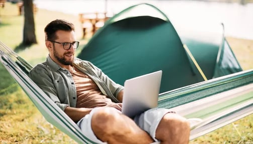 A man relaxes in a hammock, working on a laptop under a sunny sky.