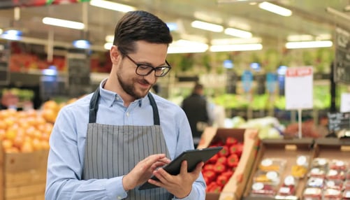 Close up portrait of happy Caucasian male worker in glasses standing in supermarket and typing on tablet.
